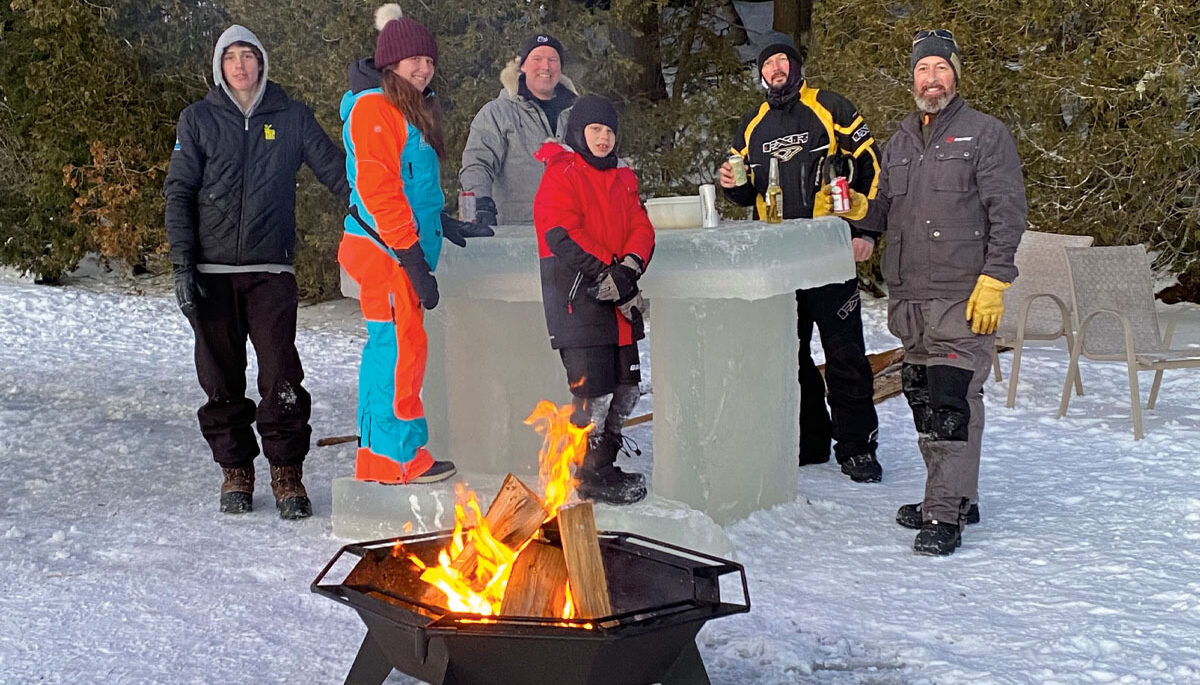 a group of people in snowsuits tand around a bar made of ice