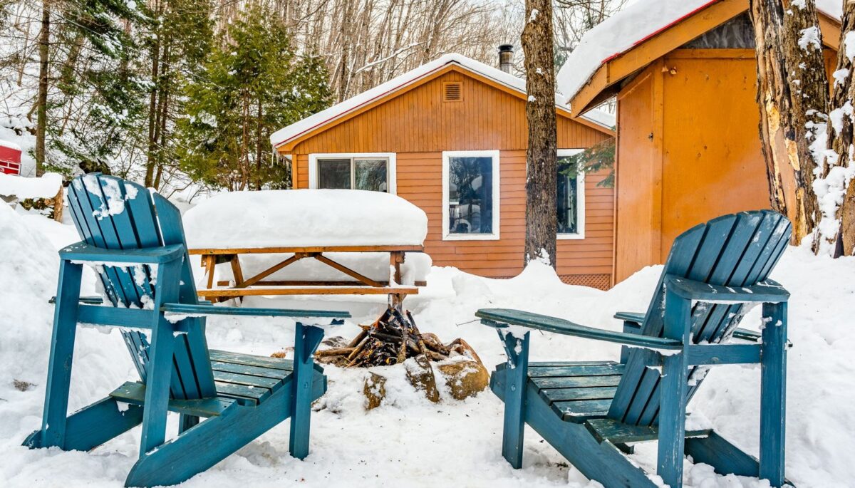 Two turquoise Muskoka chairs face a fire pit. Behind, a wood outhouse and a wood cabin