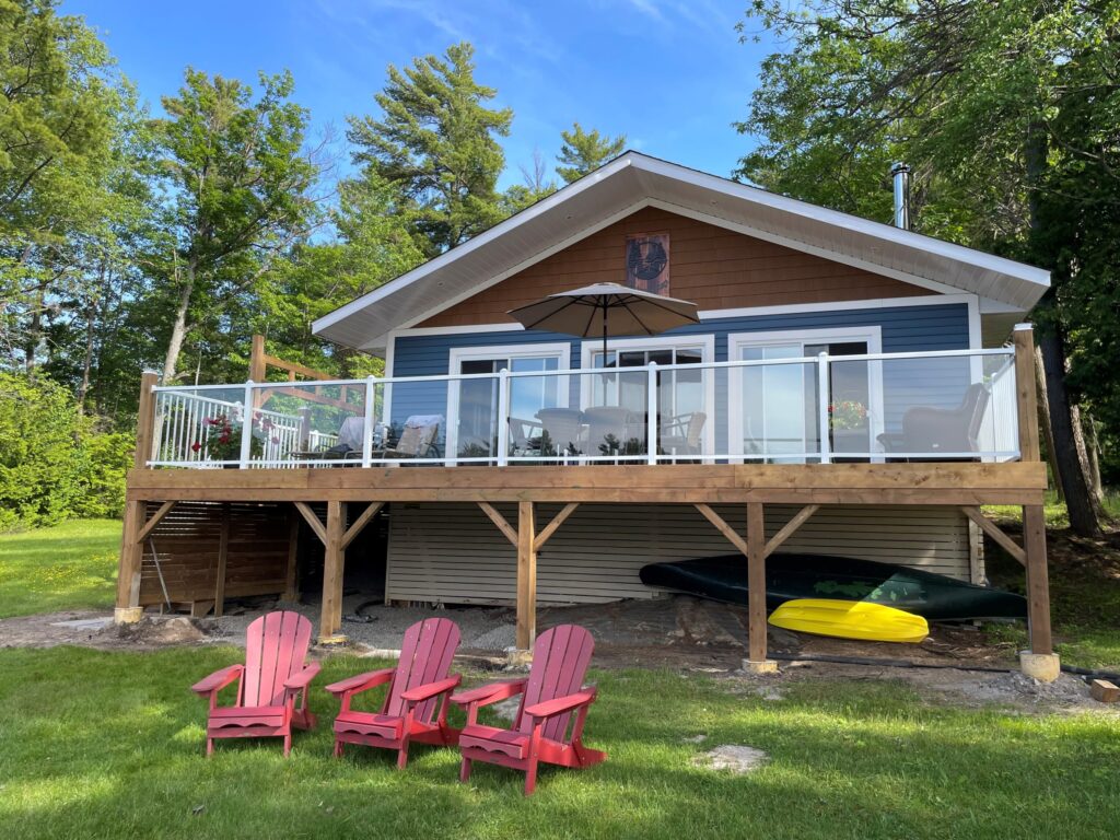 A blue-paneled cottage with a glass-framed deck on a grassy lawn. Three red Muskoka chairs sit in front