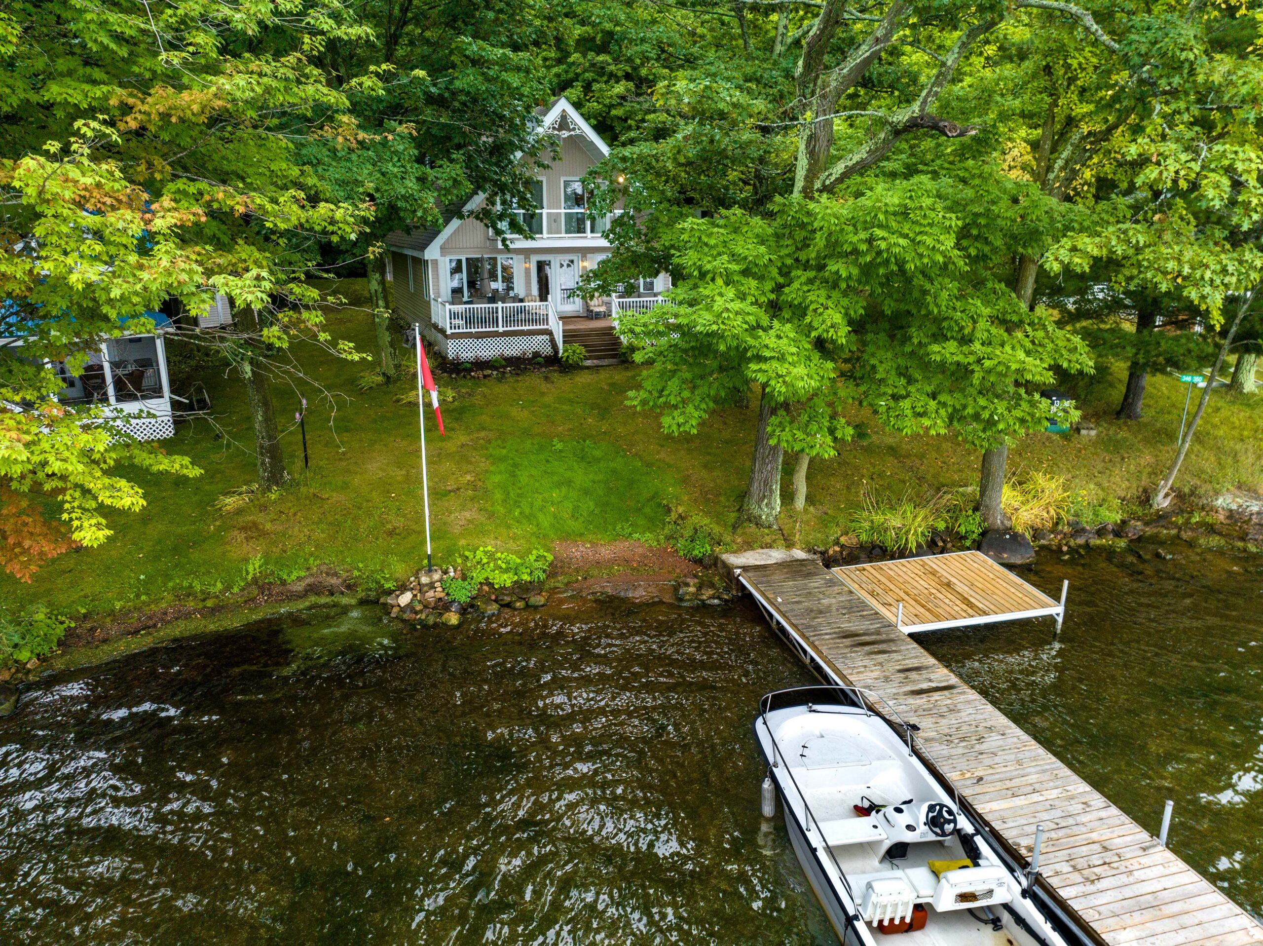 A triangular cottage on a grassy lawn. In front, a lake with a dock and a white boat parked on the left.