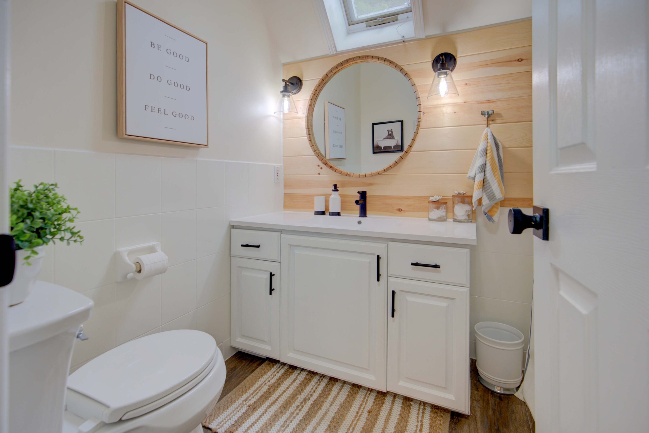 A white bathroom with white wood cabinets. A round mirror hangs above the sink on a fake wood wall