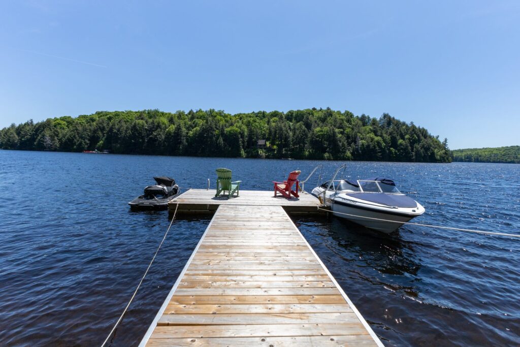A wooden dock in the water across from a forested island. At the end of the dock, two Muskoka chairs face out and a boat is parked on the right.