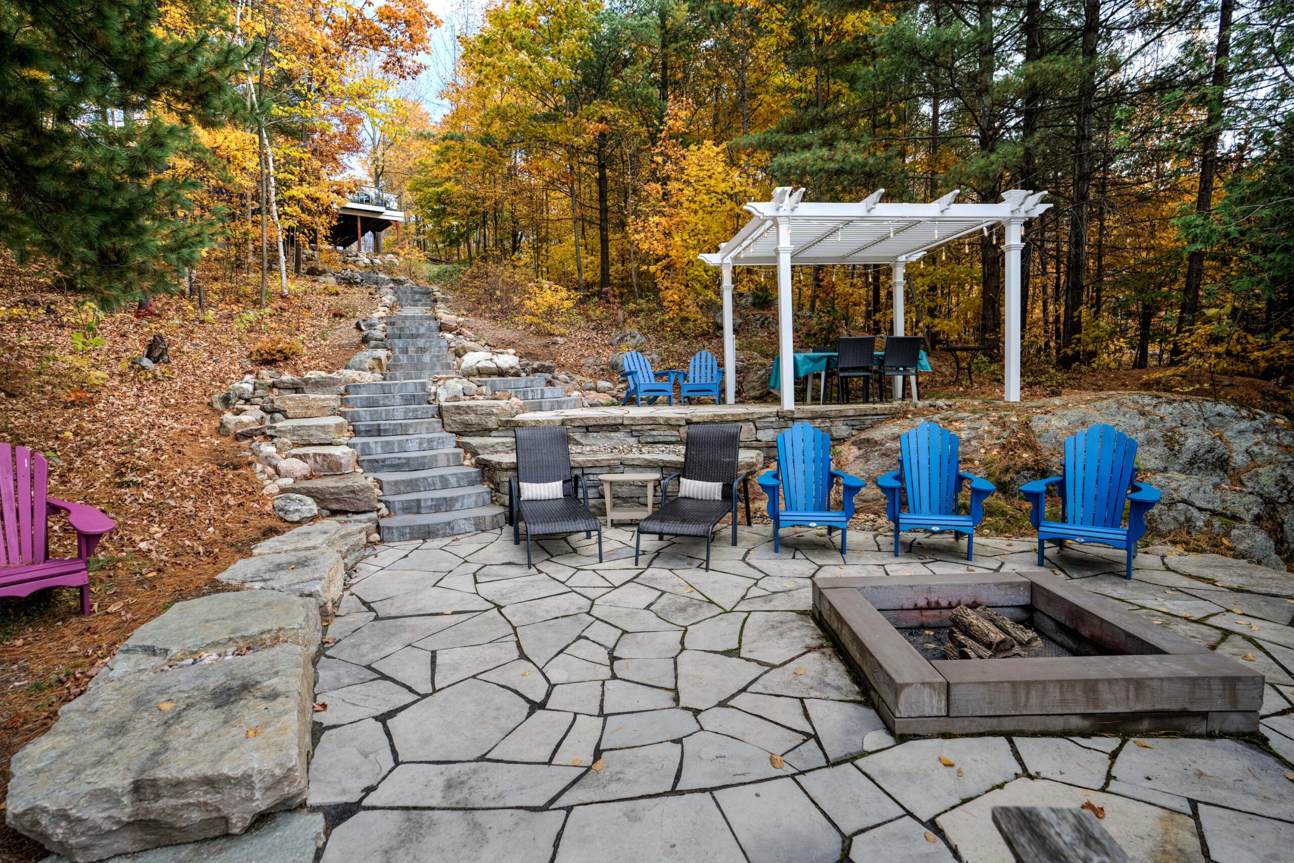 A stone patio with blue Muskoka chairs, a white pergola and fire pit