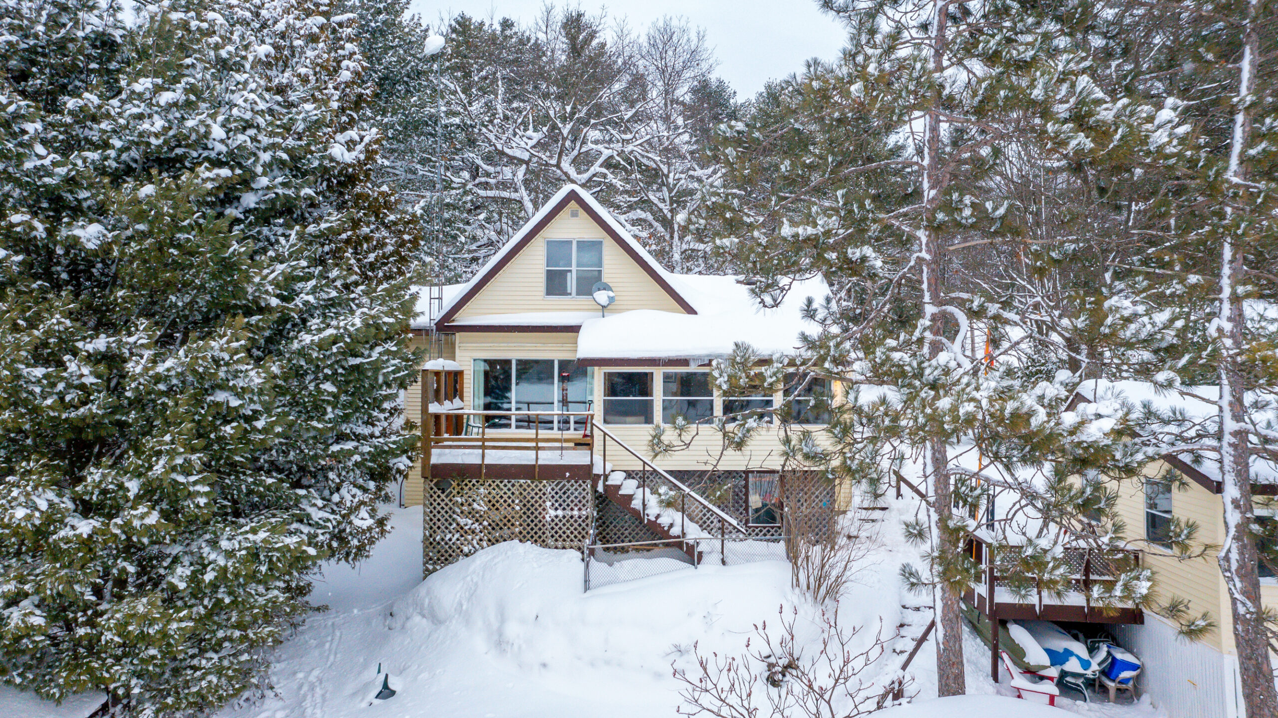 A charming, snow-covered cottage nestled among tall evergreen trees on Morrison Lake, showcasing a classic A-frame design and a wraparound deck.