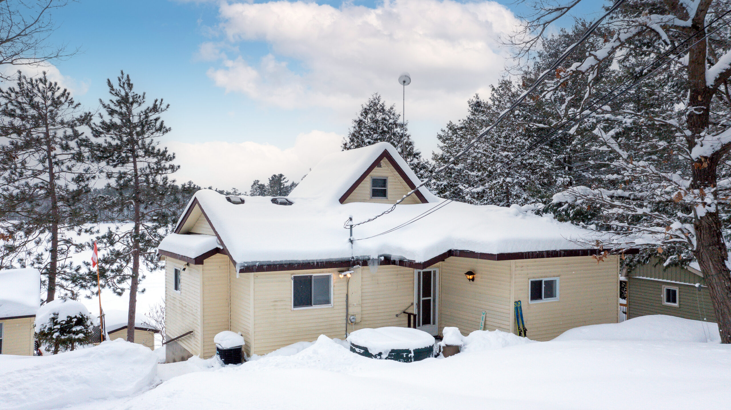 A serene winter scene featuring a lakeside cottage with a snow-covered roof, surrounded by pine trees, with glimpses of the frozen lake in the background.