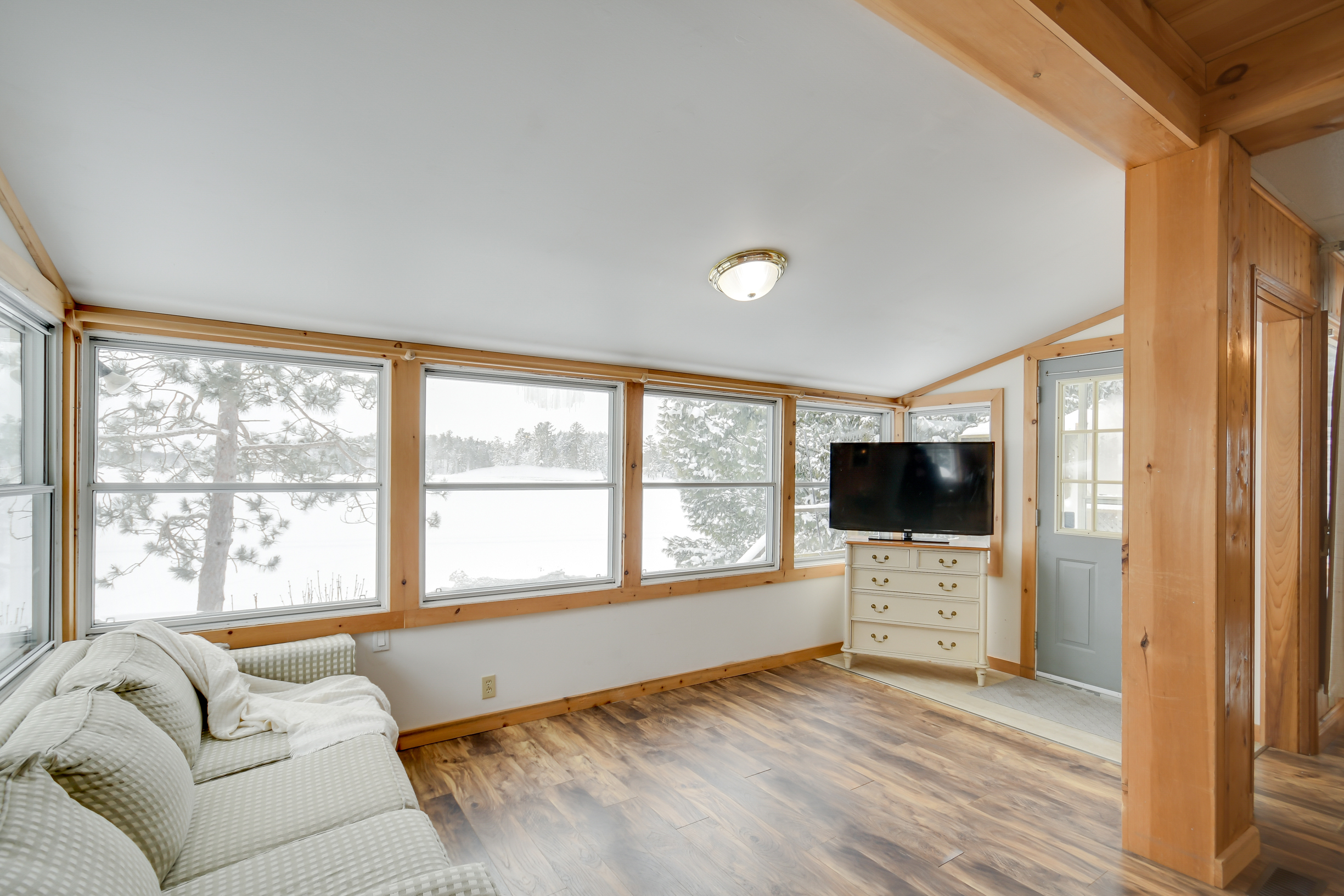 A cozy sunroom featuring large windows overlooking a snow-covered Morrison Lake, with comfortable seating and a TV for relaxation.