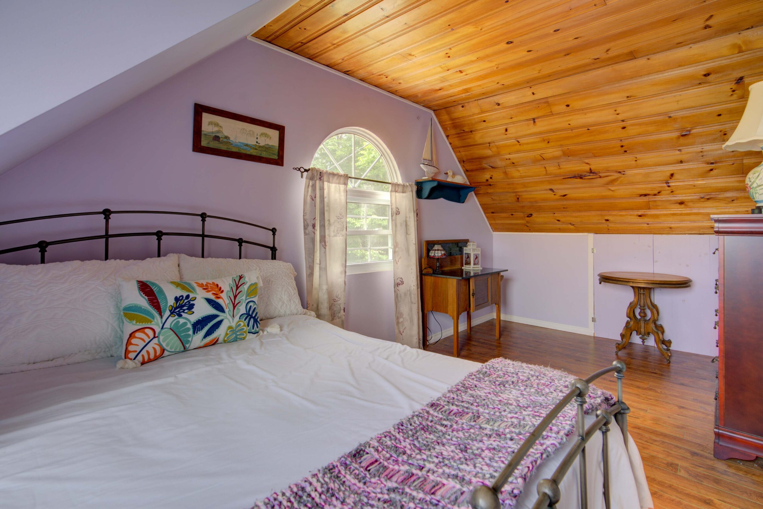 A white bed with a purple bedspread in an attic room with wood ceilings