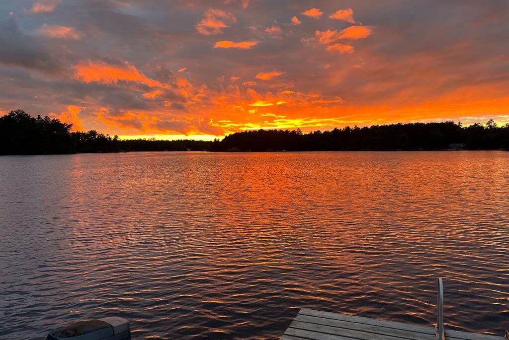 Fiery orange and gold sunset reflecting over Kahshe Lake, viewed from a dock with a boat motor in the foreground