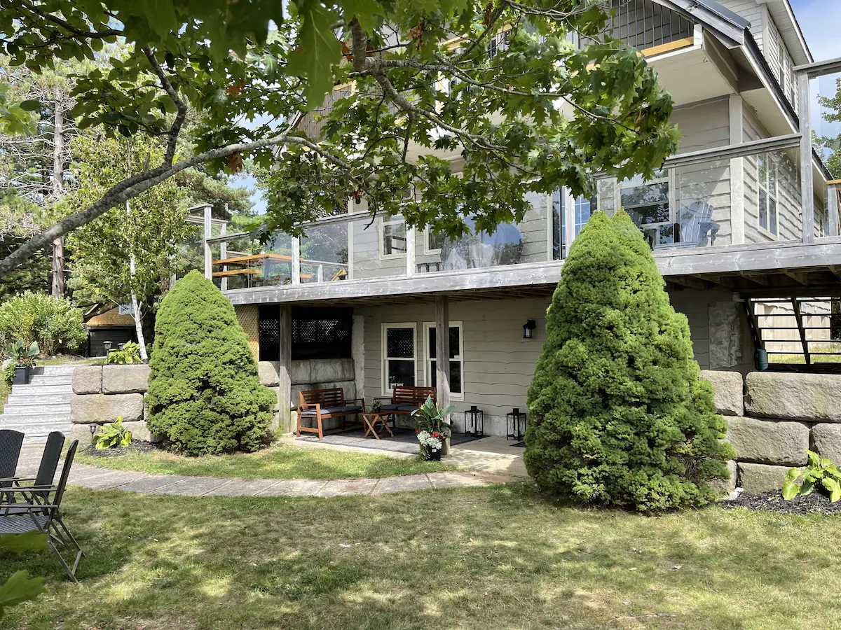 A beige cottage with an upper glass-railed deck in a green yard