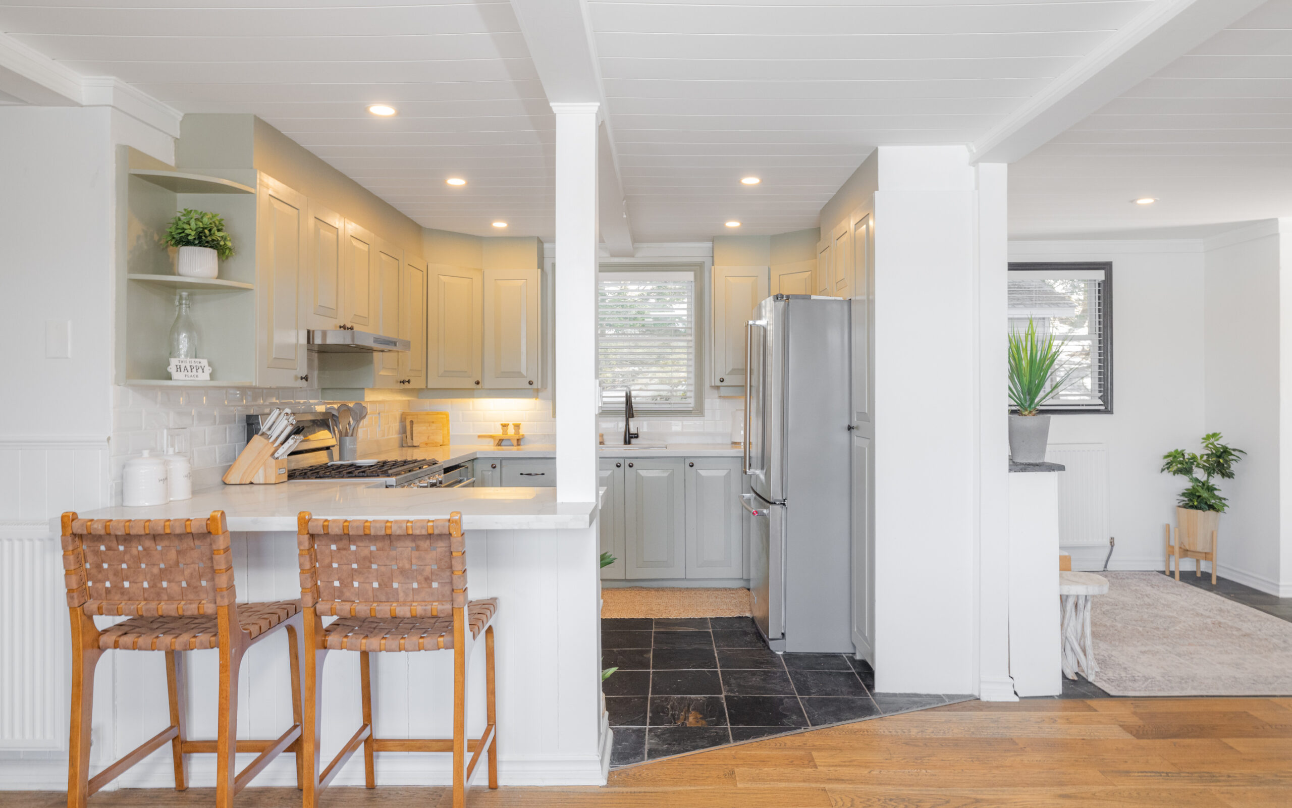 A white breakfast island with two wicker high-top chairs face a kitchen with grey-blue cabinets