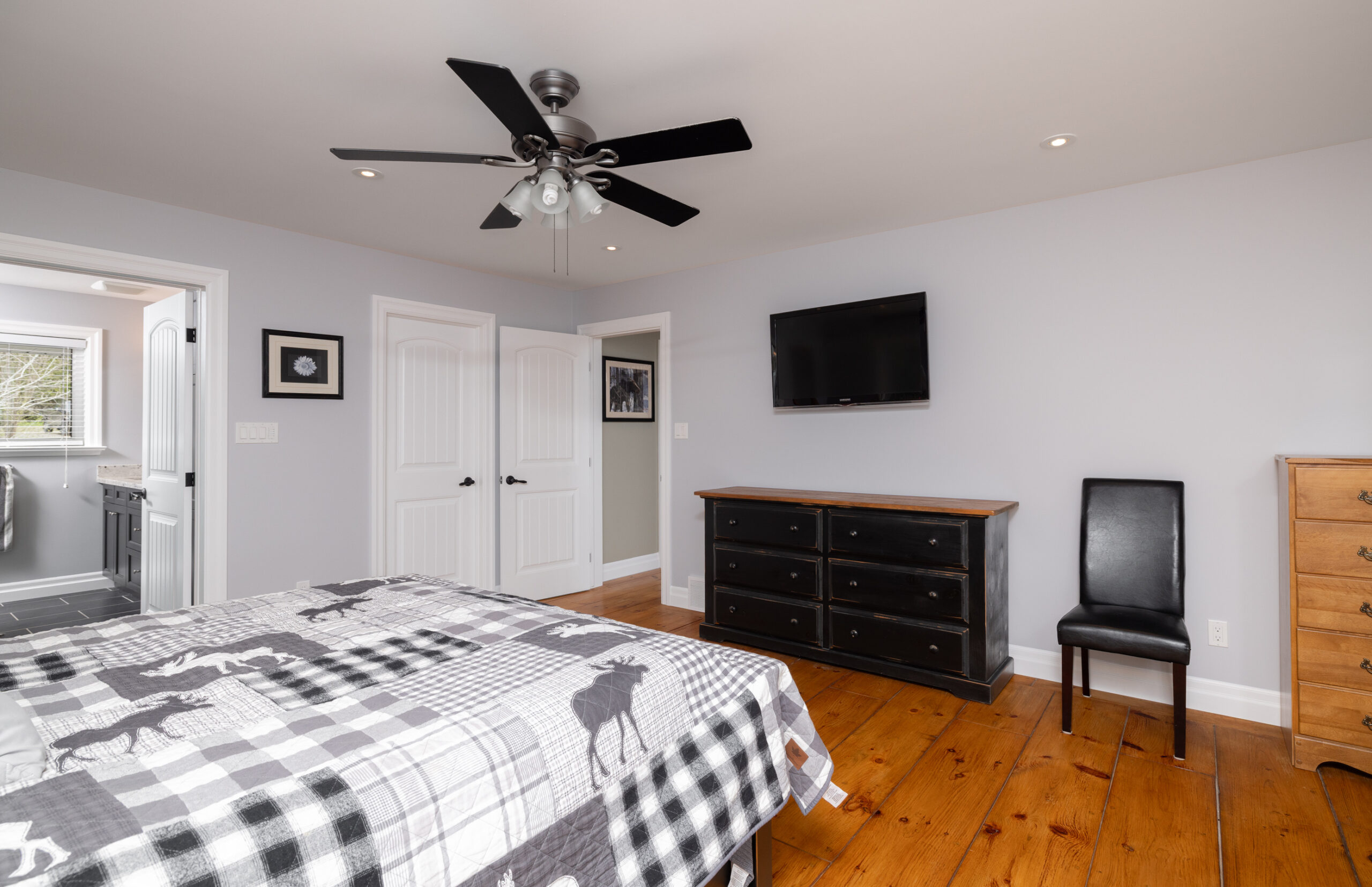 Bed in a bedroom with grey walls, hardwood floors, a dresser, and a mounted television.