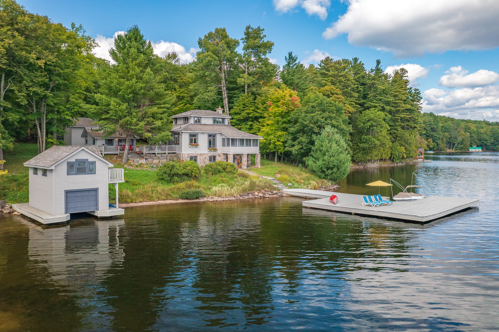 Scenic waterfront view of a Lake of Bays cottage with a two-storey boathouse, dock, and lush surrounding forest under a blue sky