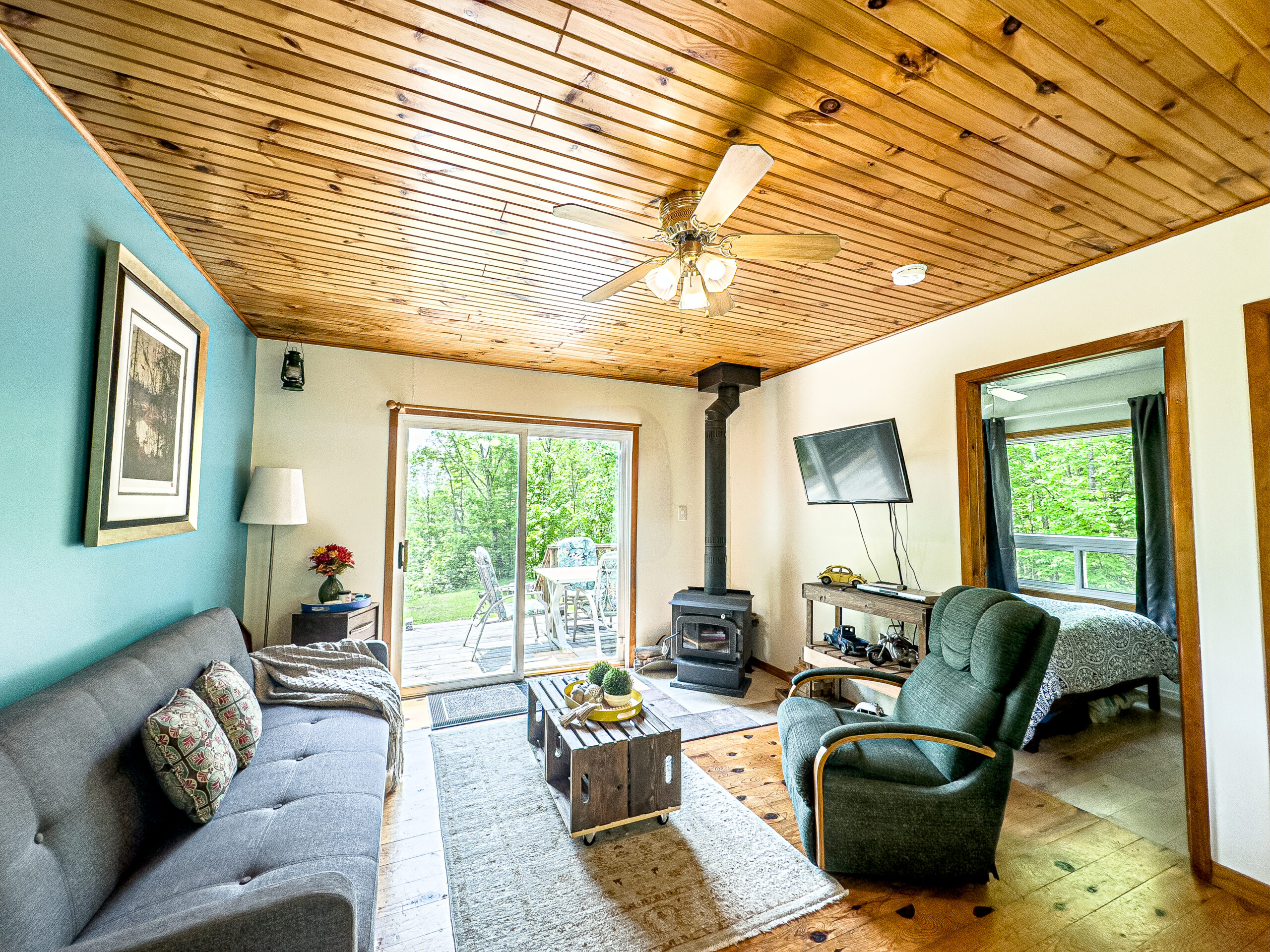 A grey couch faces a reclining chair and a wood stove in a white cottage living room. To the left, glass sliding doors lead to the deck
