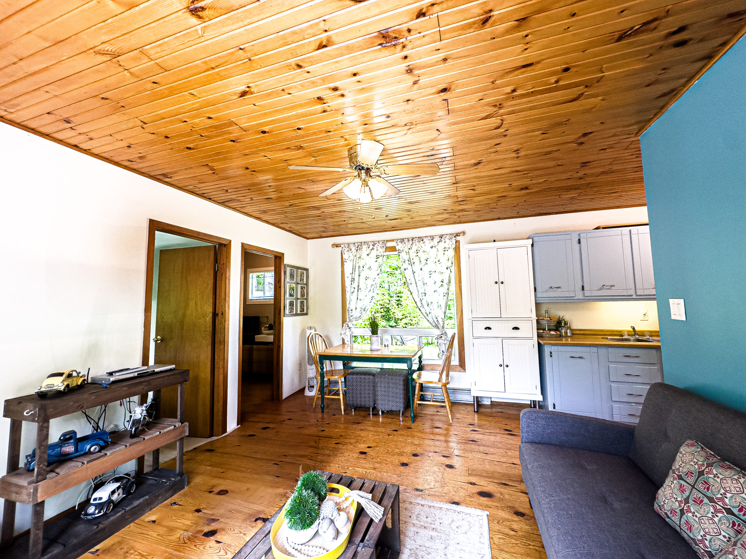 A white living room with a wood ceiling. A grey couch faces a recliner chair. Behind, a small breakfast nook next to a window