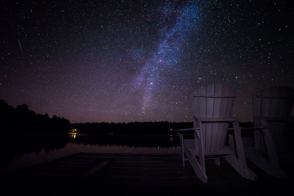 Muskoka chairs on a dock at night with the Milky Way in the background. Light pollution