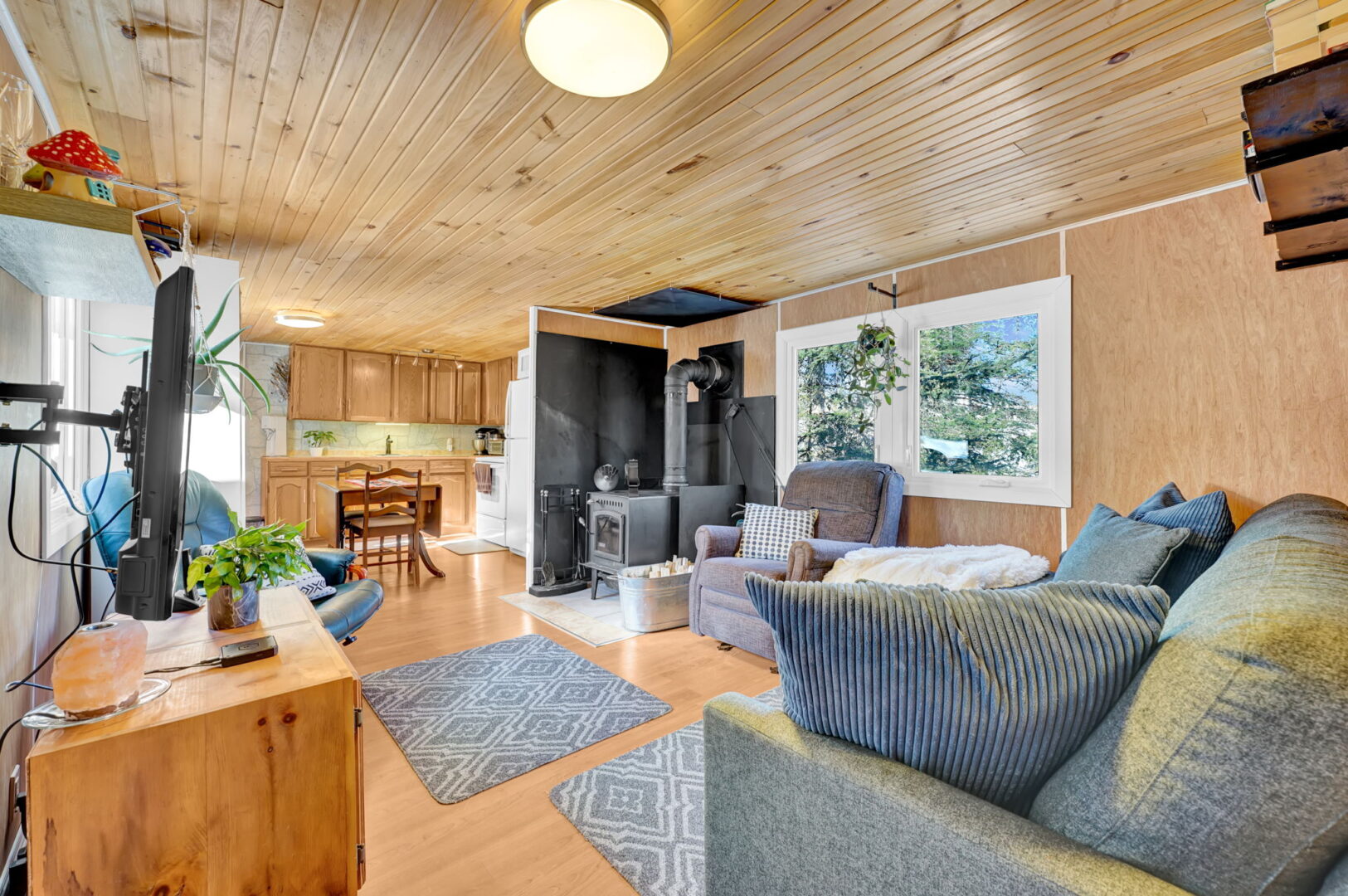 An open-concept wood-paneled living room with grey couches, a wood stove, and a view to the wood kitchen