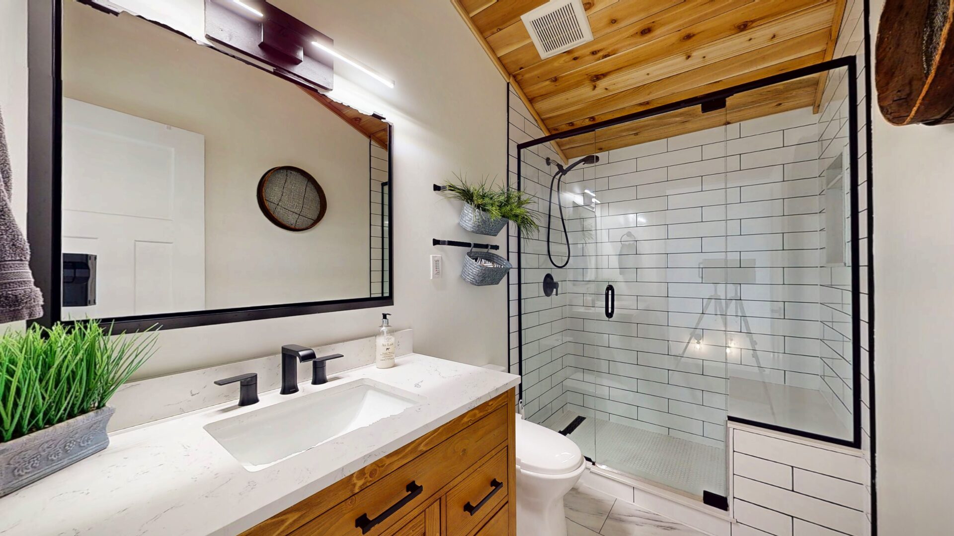 A white bathroom with a wood vanity and a black-rimmed mirror. To the right, a black and white tiled shower