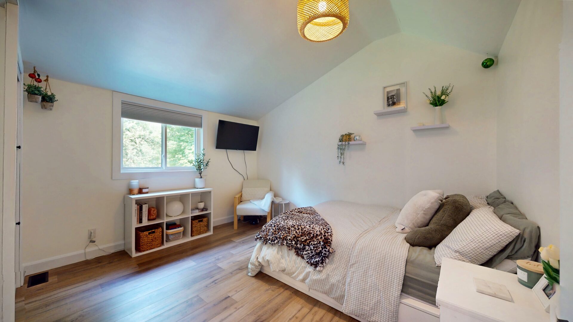 A white bed with a patterned blanket at the end facing a window and shelf