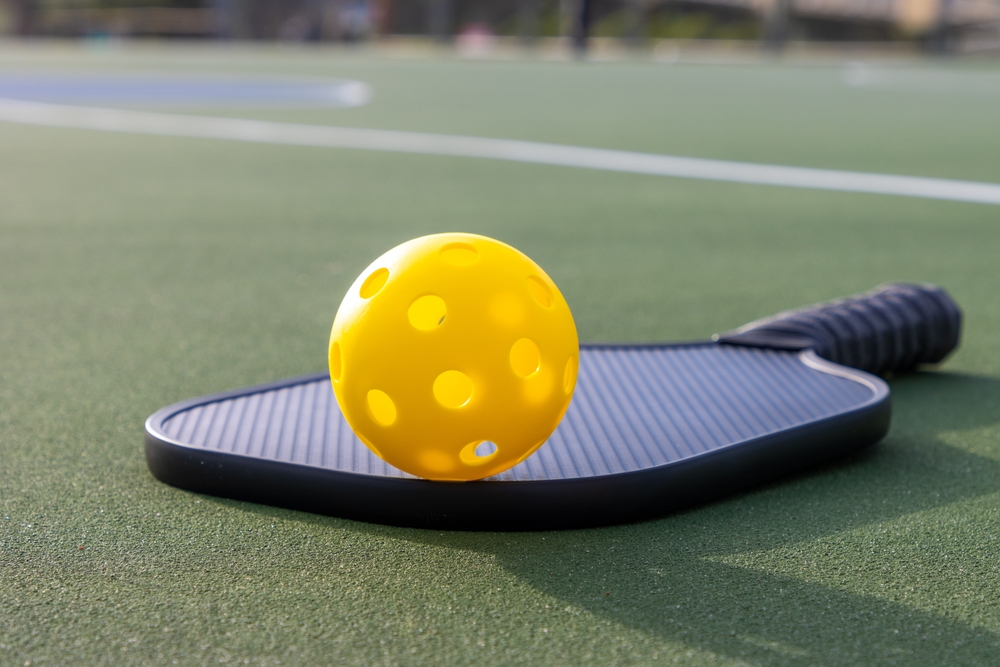 Yellow plastic pickleball ball and a black paddle on an outdoor playing court.