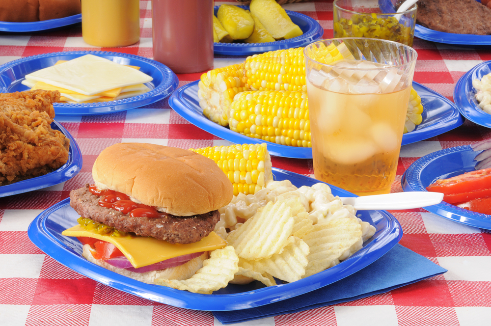 A cheeseburger with potato chips and corn on the cob on a picnic table loaded with food