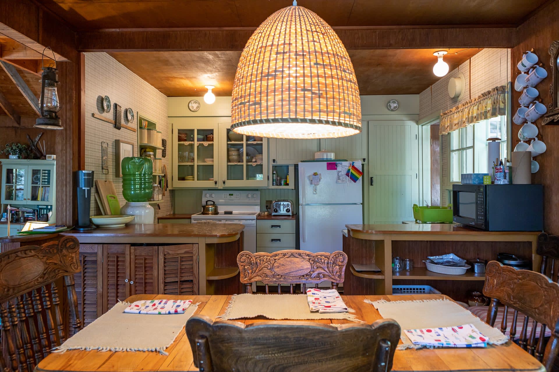 A dining table with a wicker pendant light hanging above. Behind, a light green kitchen