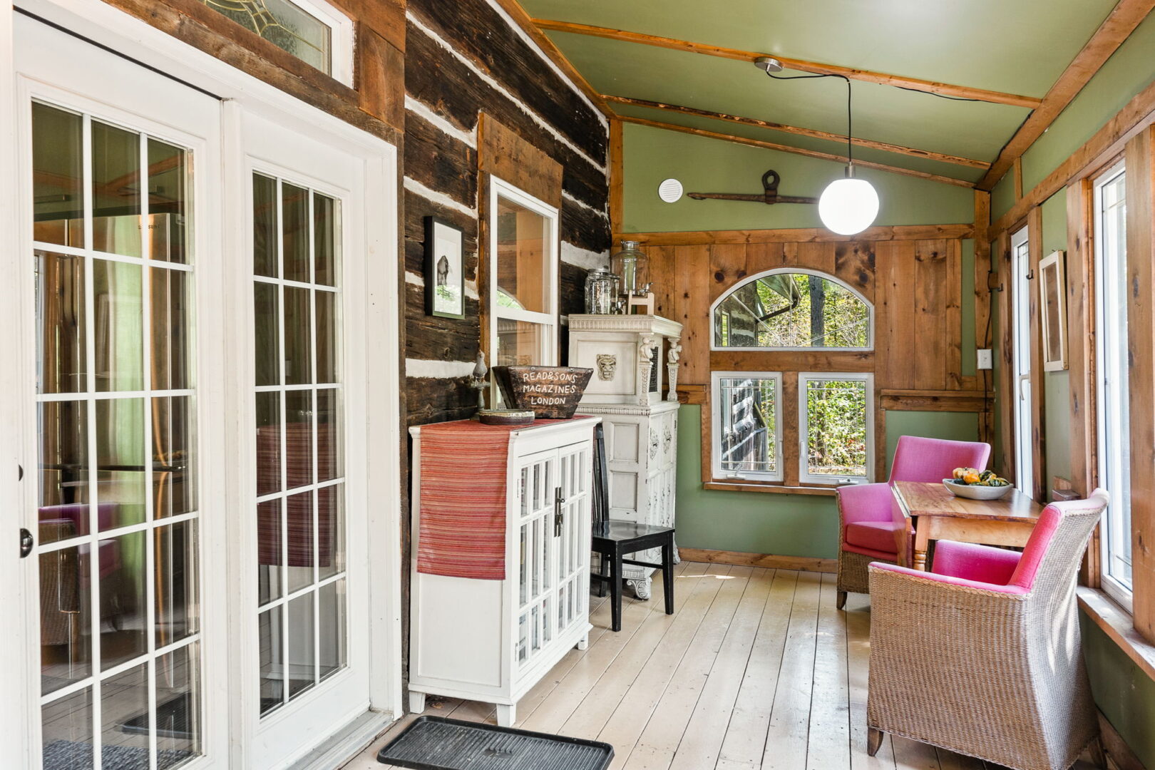Enclosed front porch with log wall, wood floors, vintage cabinets, and cozy seating overlooking the trees