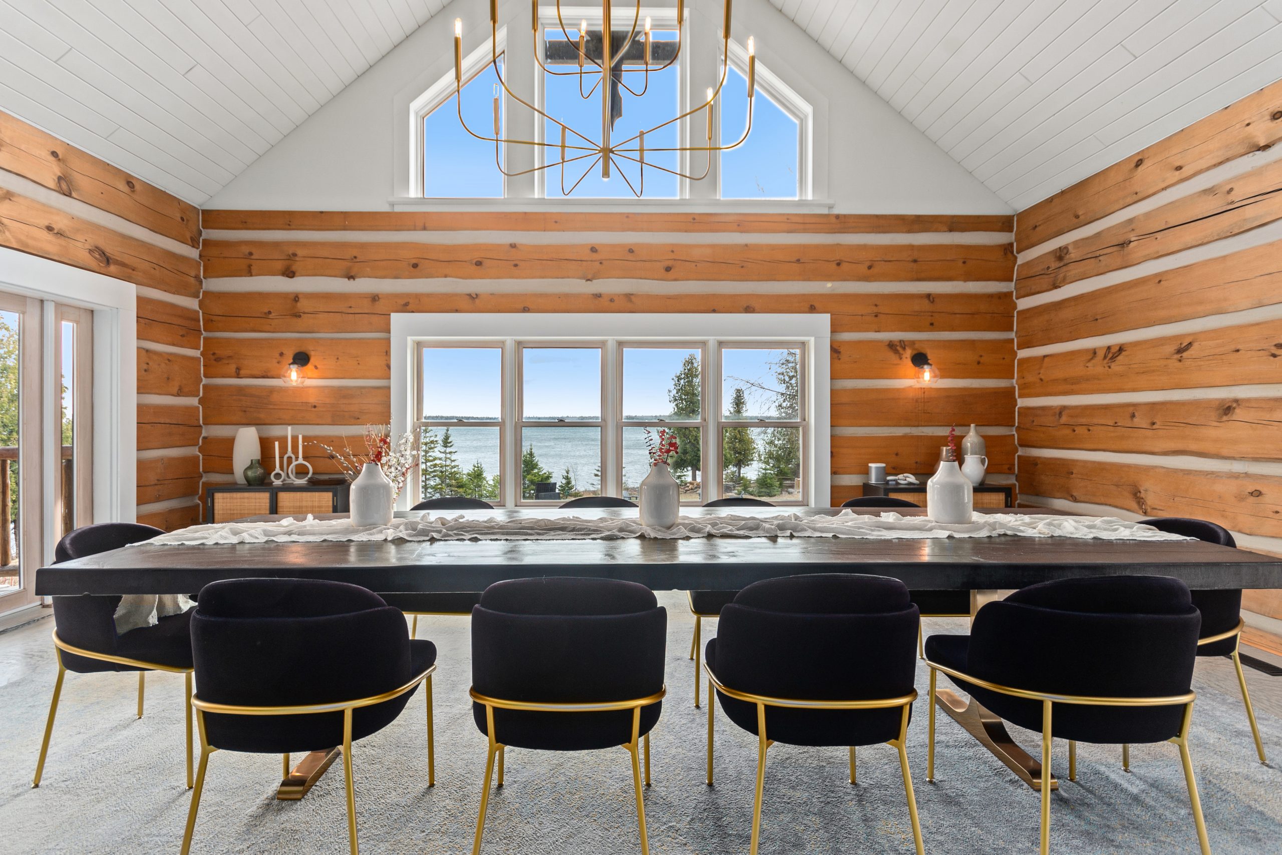 Dining space with a long table and modern chairs, set against log walls with large windows looking out to the trees.