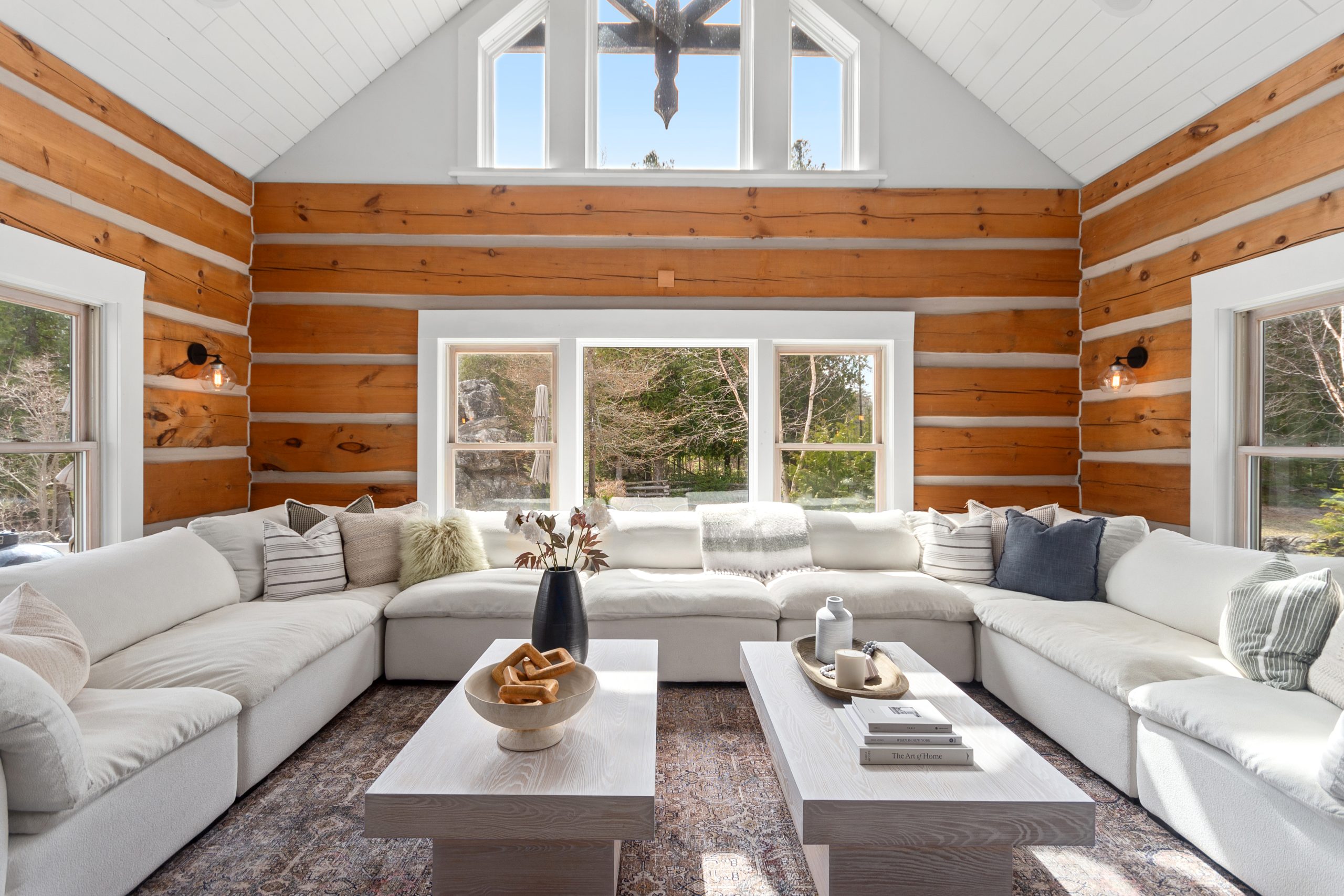 Bright great room with log walls, a vaulted ceiling, and a large white sectional facing wide windows overlooking the trees.
