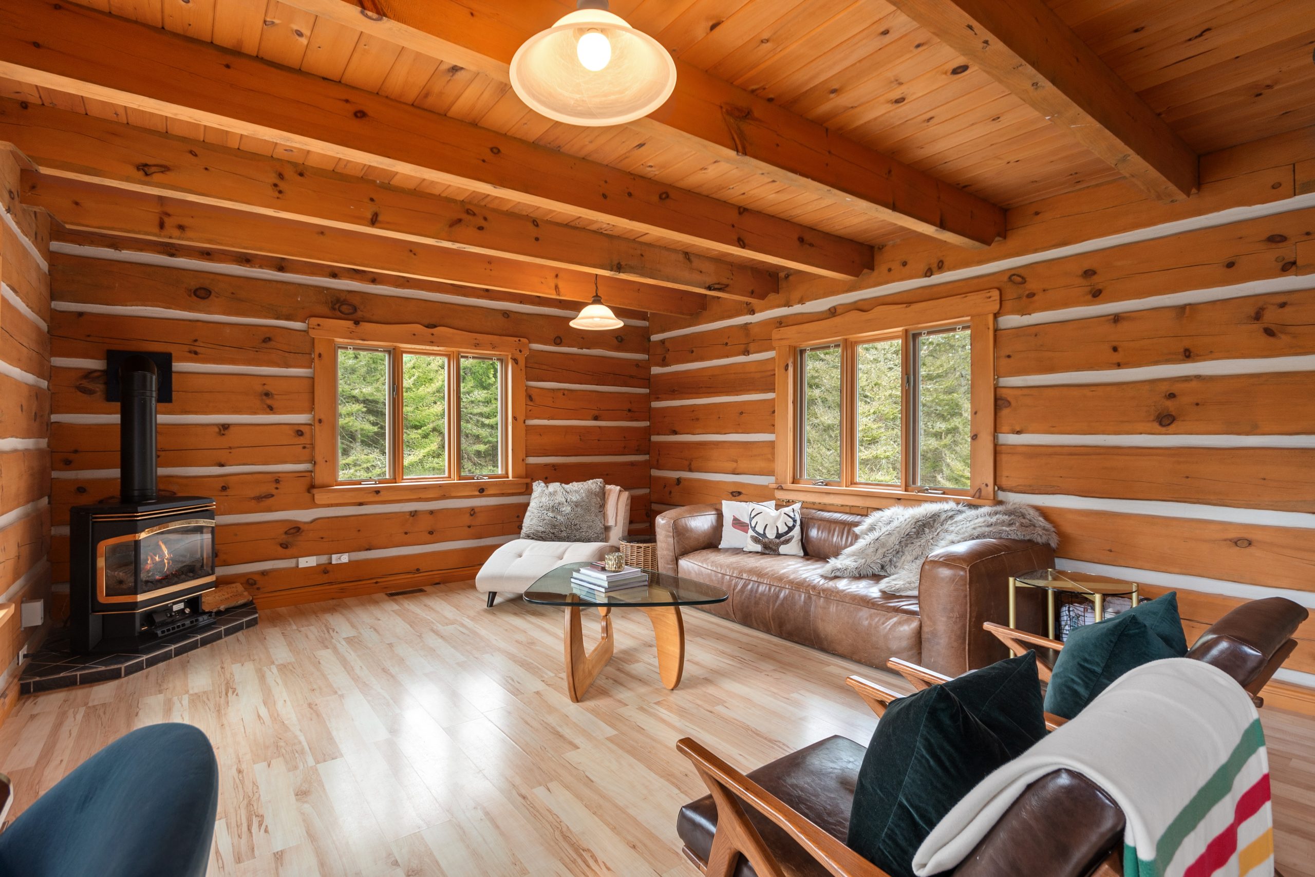Cozy living room with log walls, exposed wood ceiling, seating area, and a woodstove near the window.