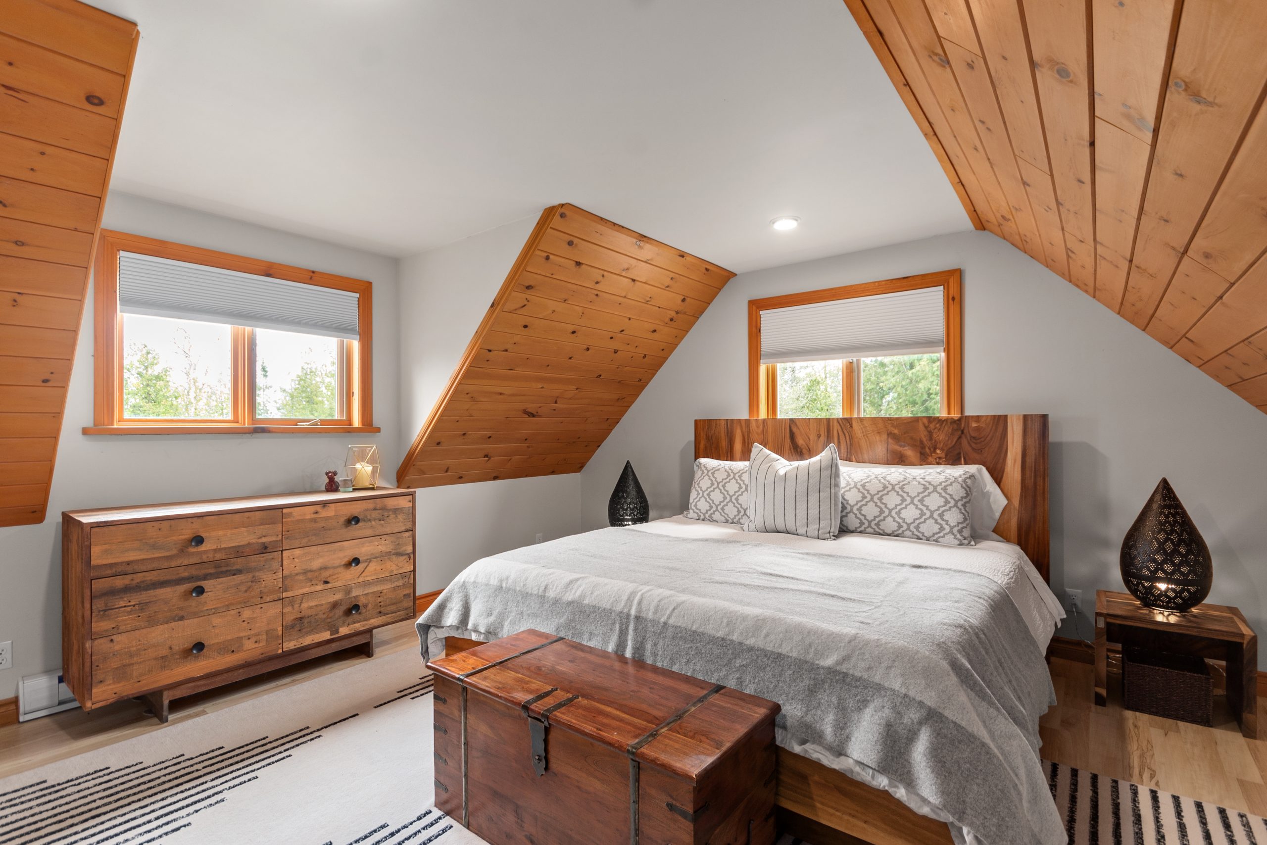 Bedroom with a bed and dresser under a sloped, vaulted ceiling with windows on either side.